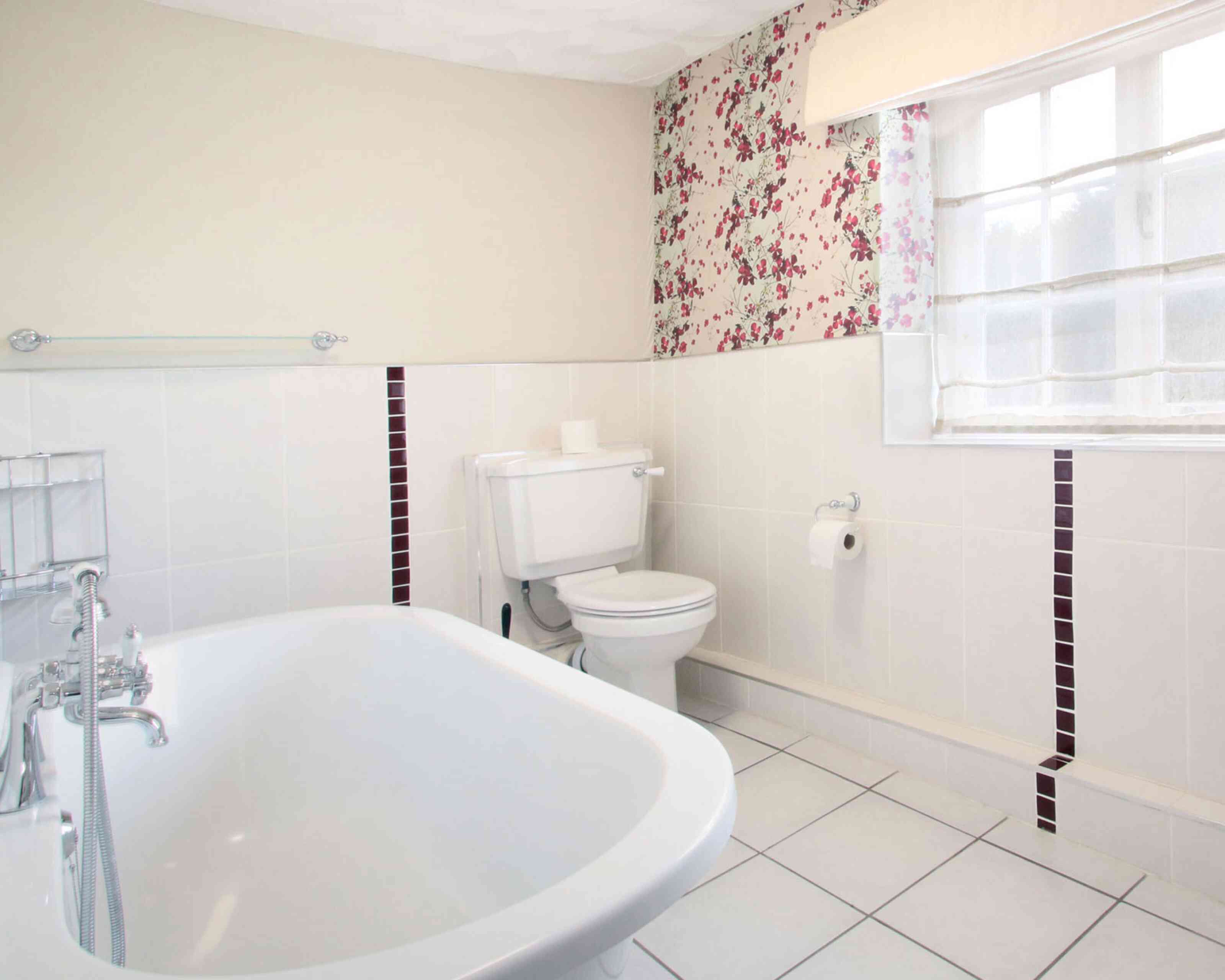 A bright and airy bathroom featuring a white bathtub, toilet, and a window with sheer curtains. The walls are decorated with floral wallpaper, and the floor is tiled in white with some dark accent tiles.