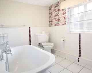 A bright and airy bathroom featuring a white bathtub, toilet, and a window with sheer curtains. The walls are decorated with floral wallpaper, and the floor is tiled in white with some dark accent tiles.