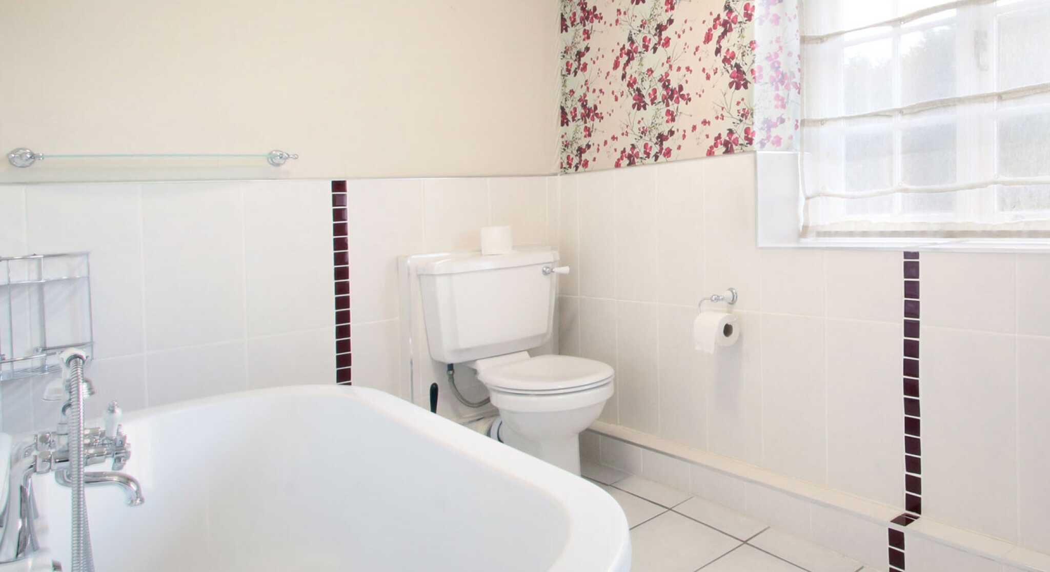 A bright and airy bathroom featuring a white bathtub, toilet, and a window with sheer curtains. The walls are decorated with floral wallpaper, and the floor is tiled in white with some dark accent tiles.
