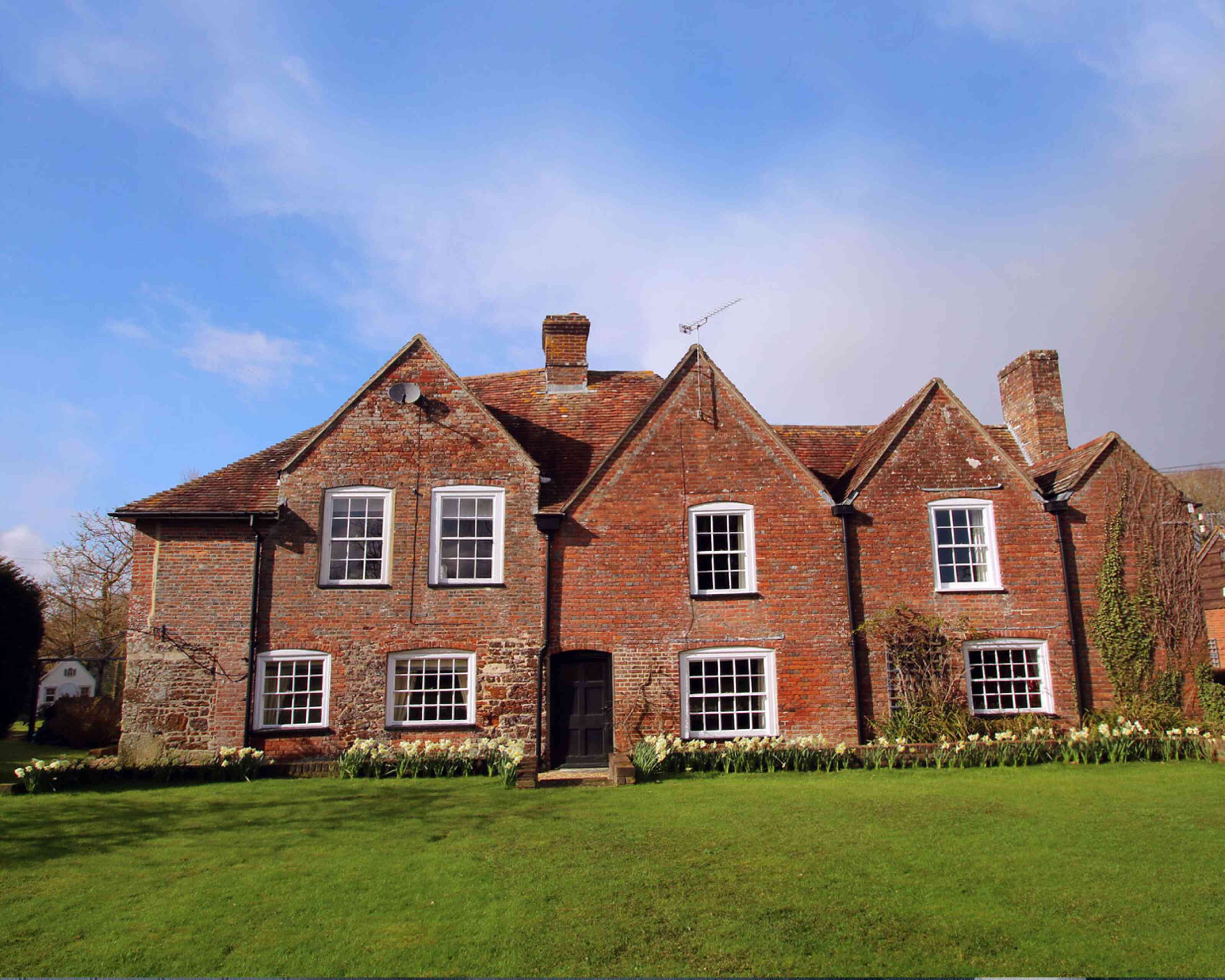 A traditional brick house with a steeply pitched roof and multiple gabled sections, set against a blue sky. The house features large windows and is surrounded by a lush green lawn adorned with blooming flowers.