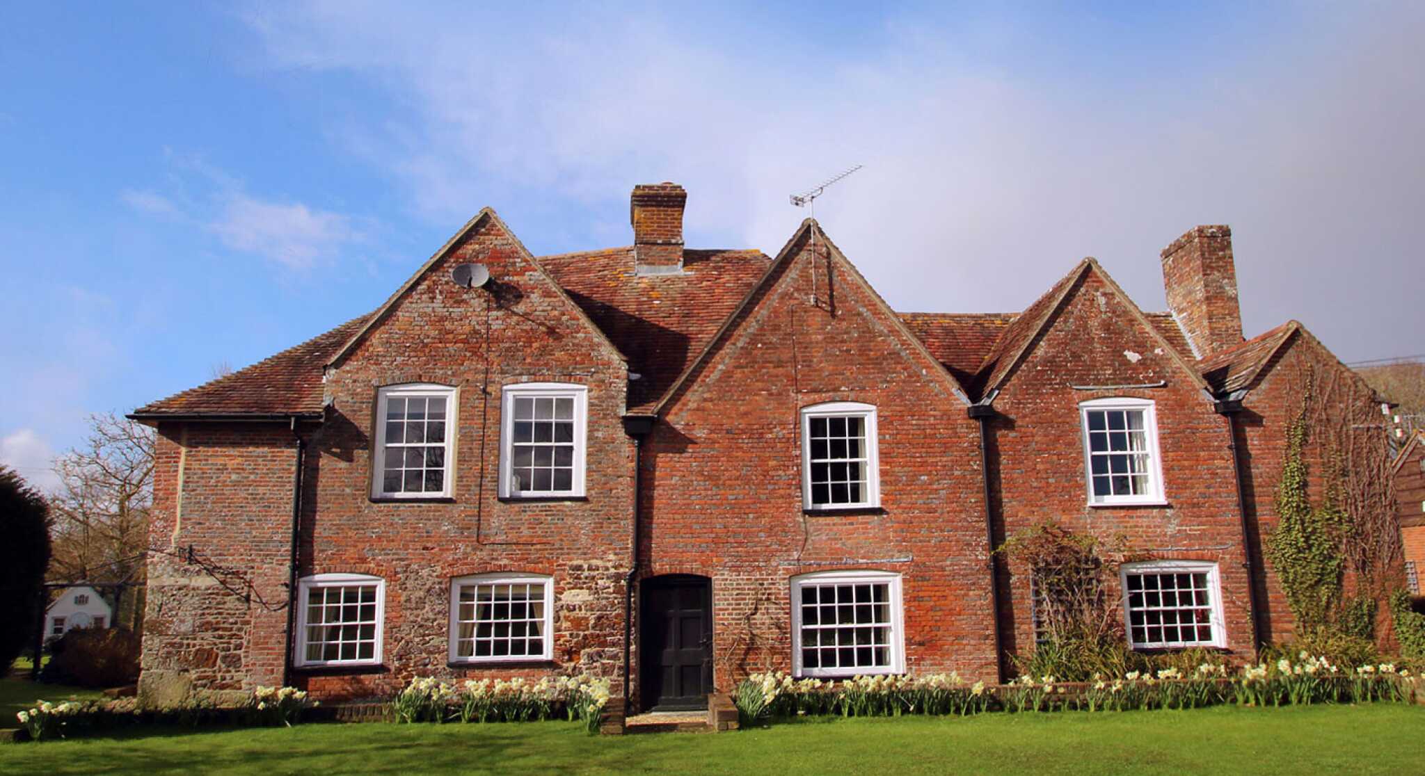 A traditional brick house with a steeply pitched roof and multiple gabled sections, set against a blue sky. The house features large windows and is surrounded by a lush green lawn adorned with blooming flowers.
