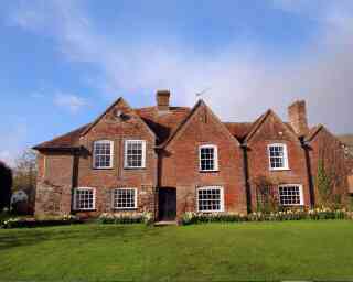 A traditional brick house with a steeply pitched roof and multiple gabled sections, set against a blue sky. The house features large windows and is surrounded by a lush green lawn adorned with blooming flowers.