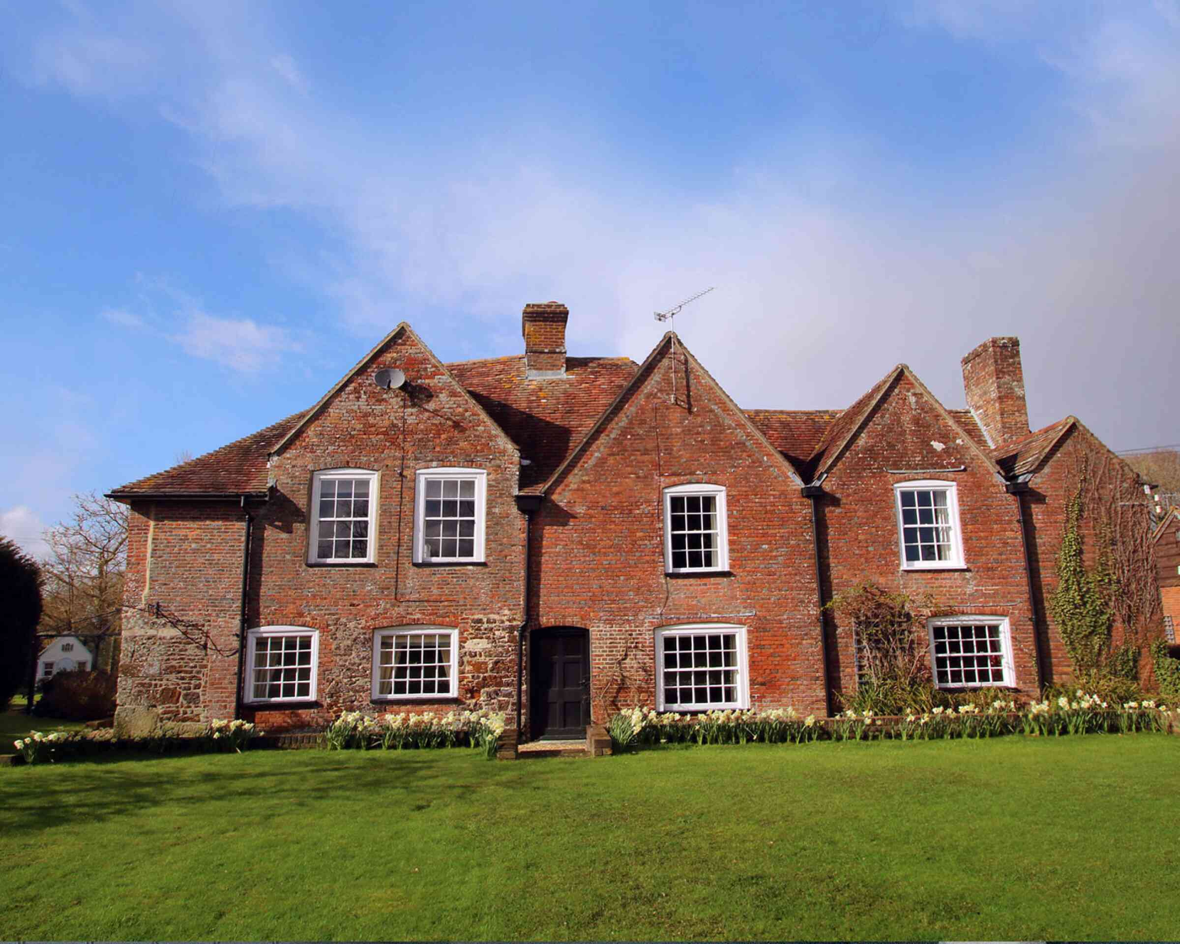 A traditional brick house with a steeply pitched roof and multiple gabled sections, set against a blue sky. The house features large windows and is surrounded by a lush green lawn adorned with blooming flowers.