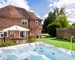 A backyard scene featuring a hot tub with jets, surrounded by a green lawn and a brick house in the background. A patio area with outdoor seating and a sunshade is visible, along with trees and a stone wall. The sky is partly cloudy.