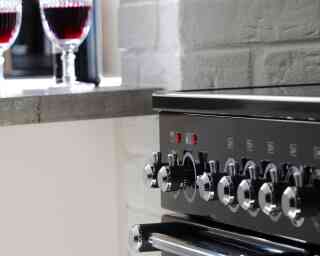 A close-up of a black oven with shiny knobs, alongside a countertop featuring two glasses of red wine.