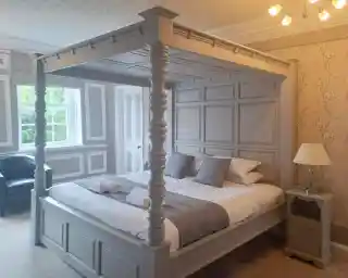A stylish bedroom featuring a gray four-poster bed with decorative paneling, crisp white linens, and gray accent pillows. A small nightstand with a lamp sits beside the bed. A plush chair is visible in the corner, and floral wallpaper adds elegance to the space.