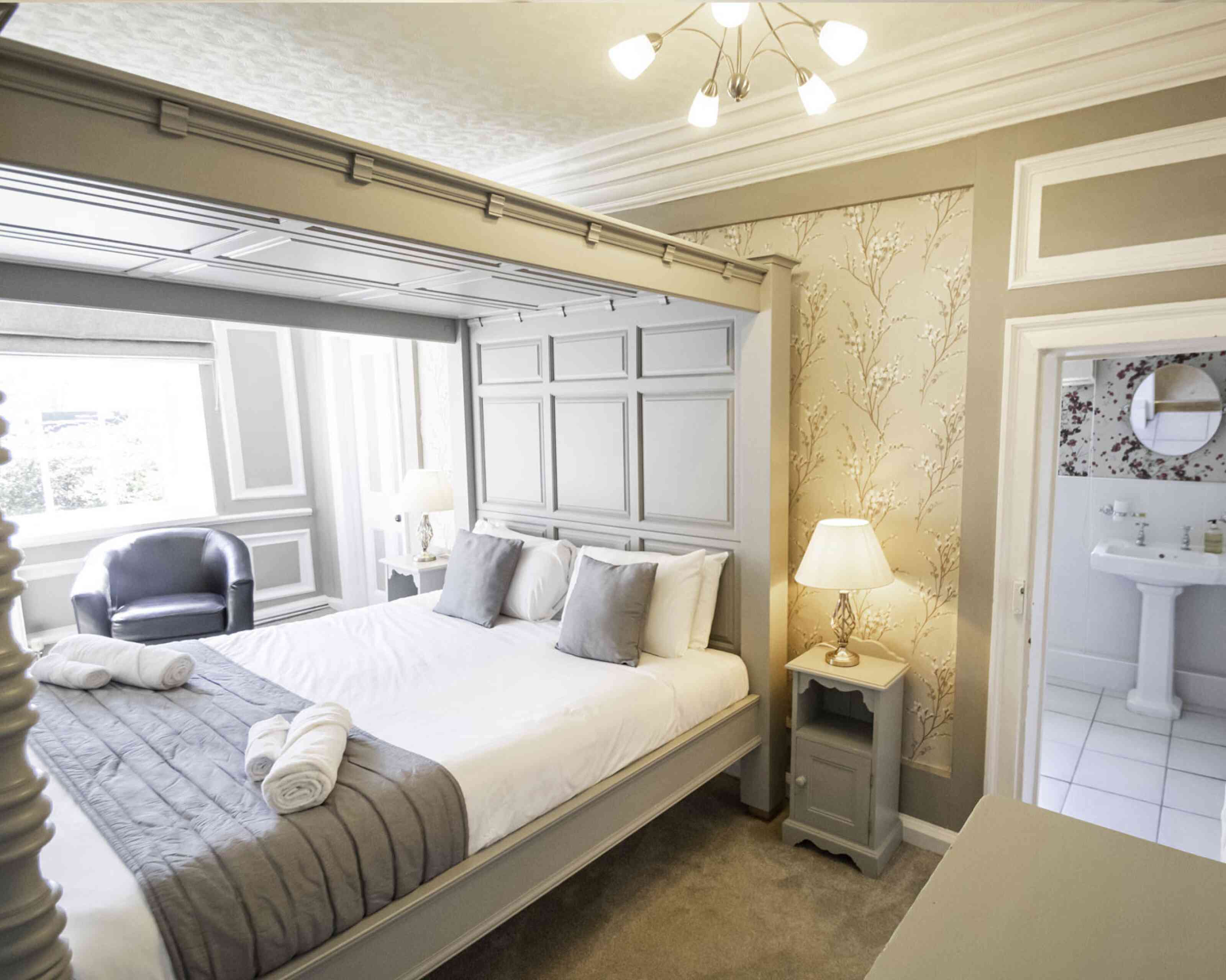 A stylish bedroom featuring a large gray bed with decorative pillows, an elegant lamp on a nightstand, a cozy armchair, and a visible en-suite bathroom. The walls are adorned with soft floral wallpaper and the room has natural light from a window.