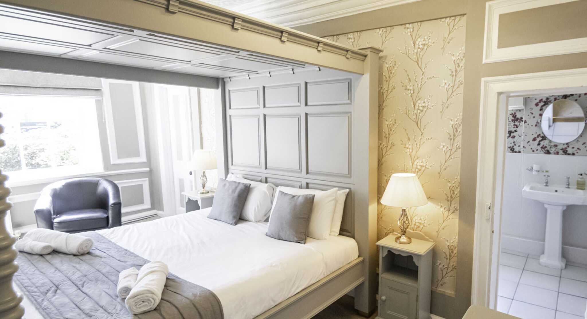A stylish bedroom featuring a large gray bed with decorative pillows, an elegant lamp on a nightstand, a cozy armchair, and a visible en-suite bathroom. The walls are adorned with soft floral wallpaper and the room has natural light from a window.