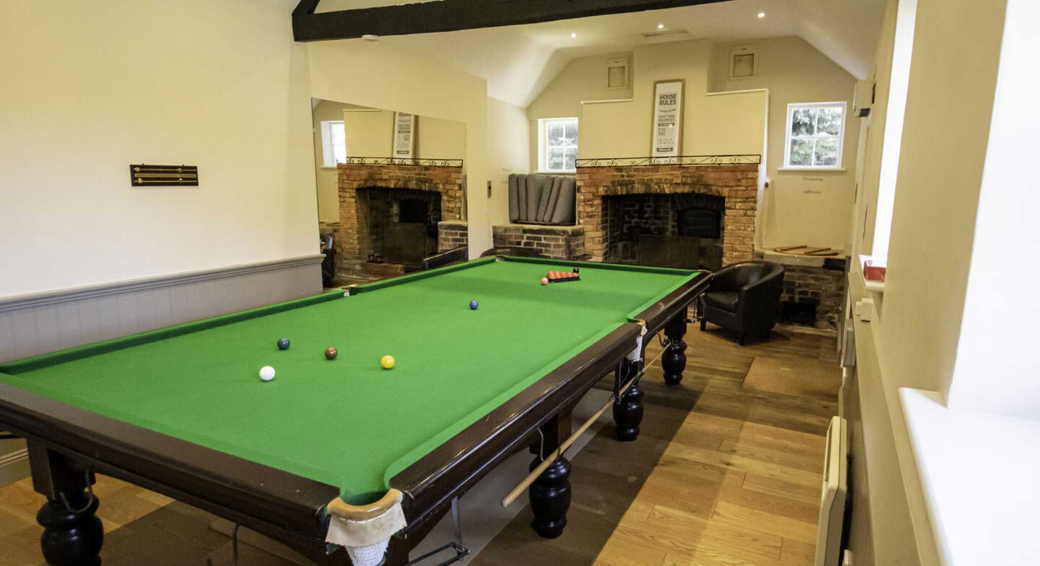 A spacious room featuring a green billiards table with balls scattered on it. Two large mirrors reflect the room's interior, which includes exposed brick walls and dark wooden beams. A comfortable chair is positioned nearby, and there are windows providing natural light.