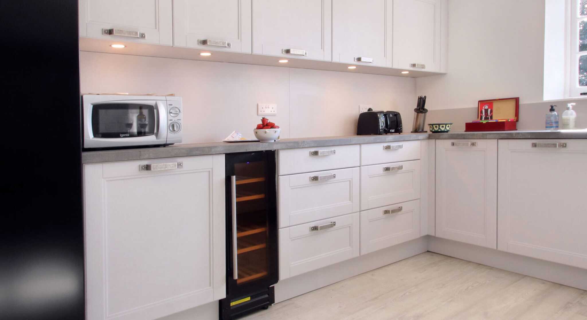 Modern kitchen featuring sleek white cabinetry, a silver microwave, black toaster, and a wine cooler. The countertop is a light gray, and the floor is a pale wood finish. Soft lighting highlights the space.