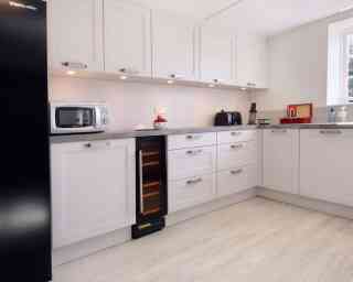 Modern kitchen featuring sleek white cabinetry, a silver microwave, black toaster, and a wine cooler. The countertop is a light gray, and the floor is a pale wood finish. Soft lighting highlights the space.