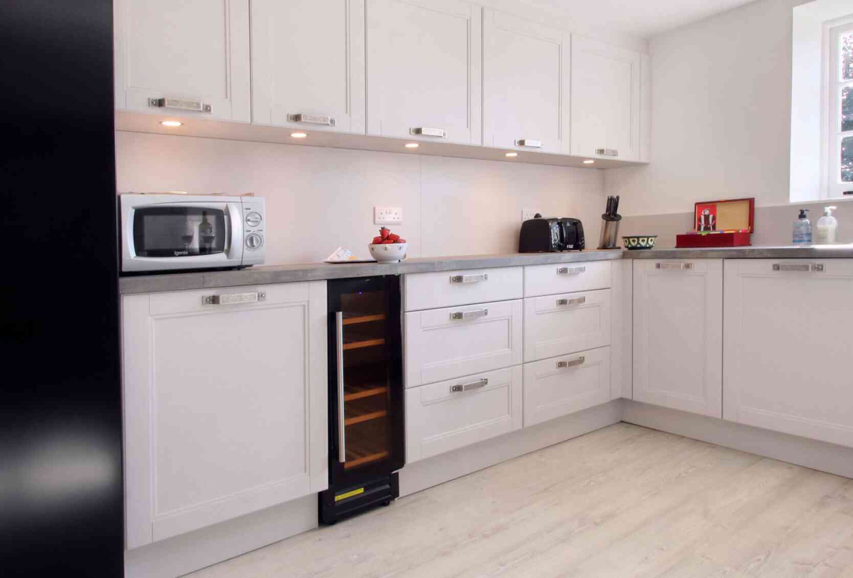 Modern kitchen featuring sleek white cabinetry, a silver microwave, black toaster, and a wine cooler. The countertop is a light gray, and the floor is a pale wood finish. Soft lighting highlights the space.