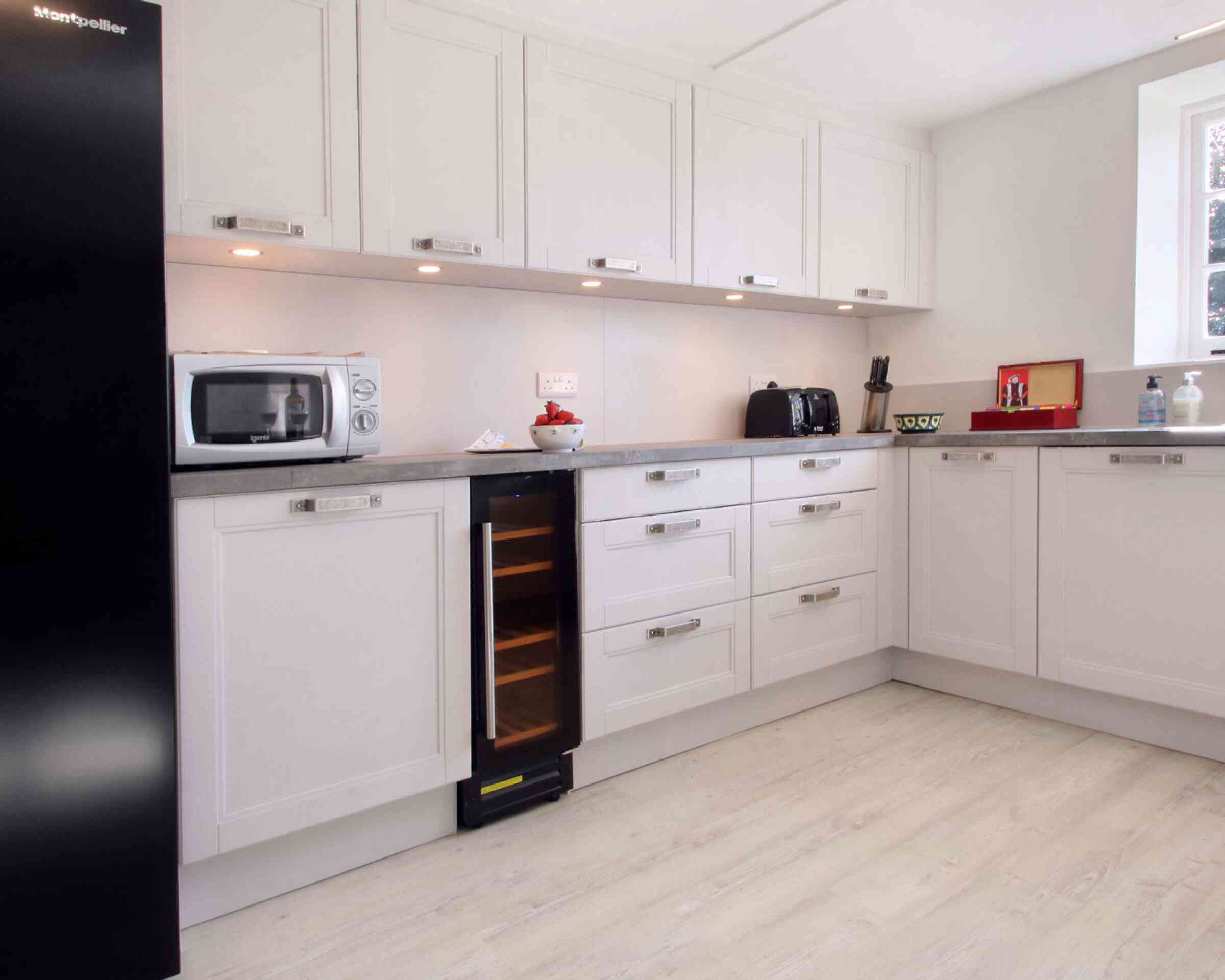 Modern kitchen featuring sleek white cabinetry, a silver microwave, black toaster, and a wine cooler. The countertop is a light gray, and the floor is a pale wood finish. Soft lighting highlights the space.