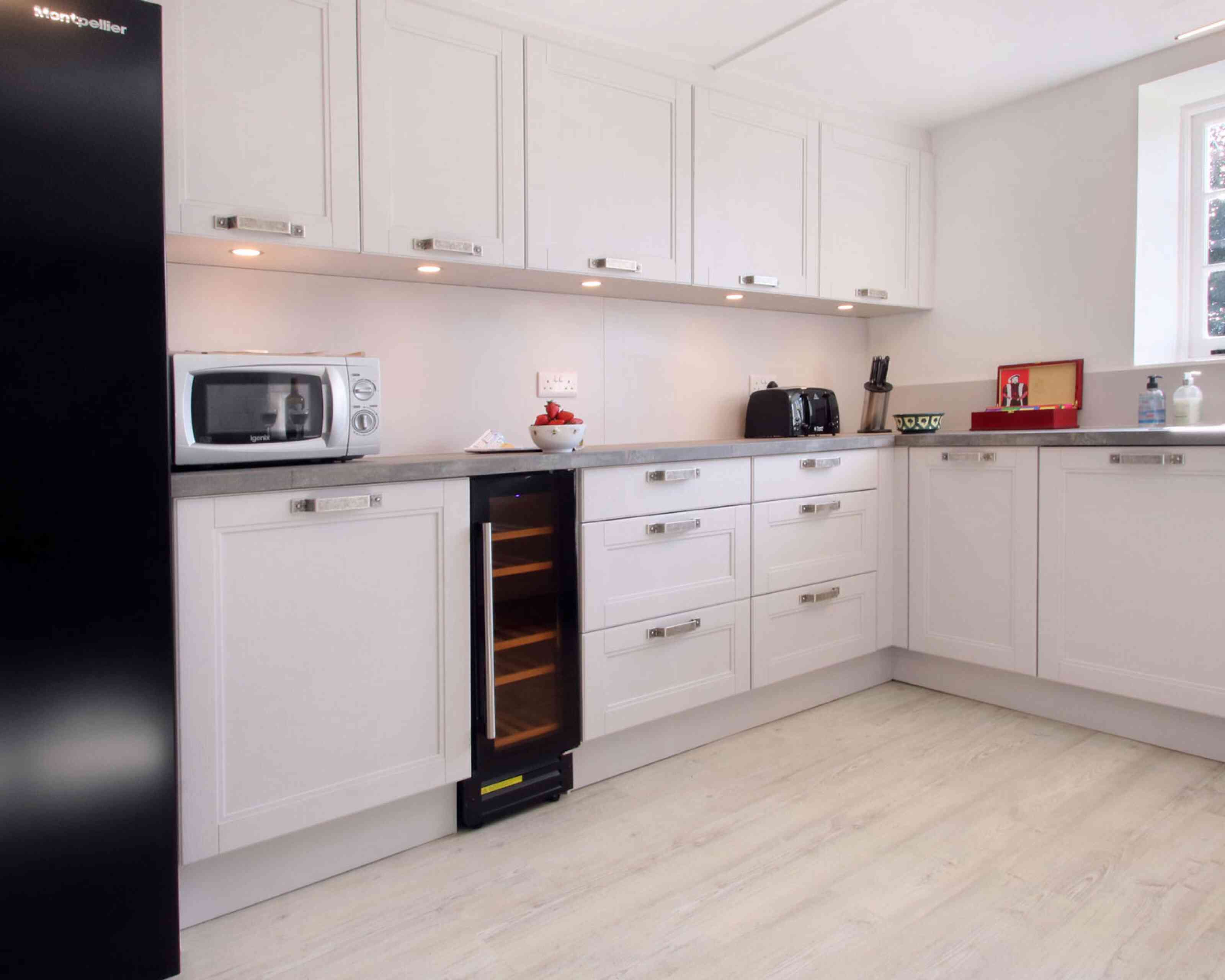 Modern kitchen featuring sleek white cabinetry, a silver microwave, black toaster, and a wine cooler. The countertop is a light gray, and the floor is a pale wood finish. Soft lighting highlights the space.