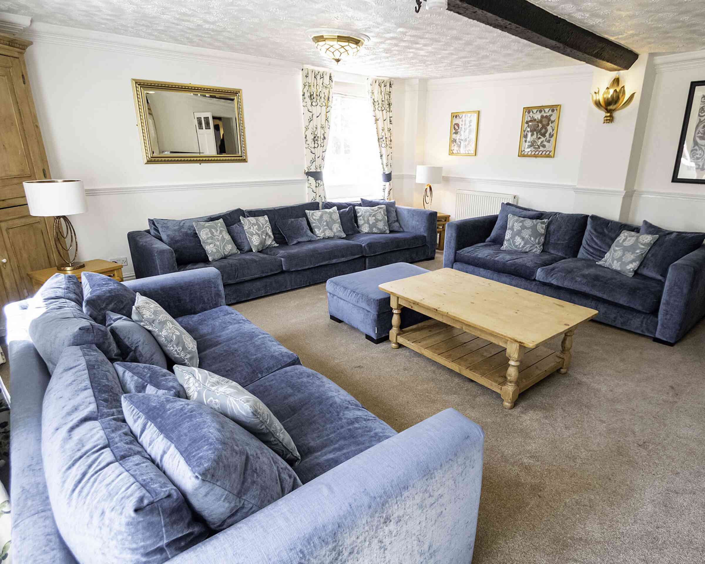 A spacious living room featuring two large blue sofas and an ottoman, all adorned with decorative cushions. There's a wooden coffee table in the center, surrounded by light-colored carpet. Wall decorations include a mirror and framed art, with natural light streaming in through large windows.