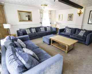 A spacious living room featuring two large blue sofas and an ottoman, all adorned with decorative cushions. There's a wooden coffee table in the center, surrounded by light-colored carpet. Wall decorations include a mirror and framed art, with natural light streaming in through large windows.