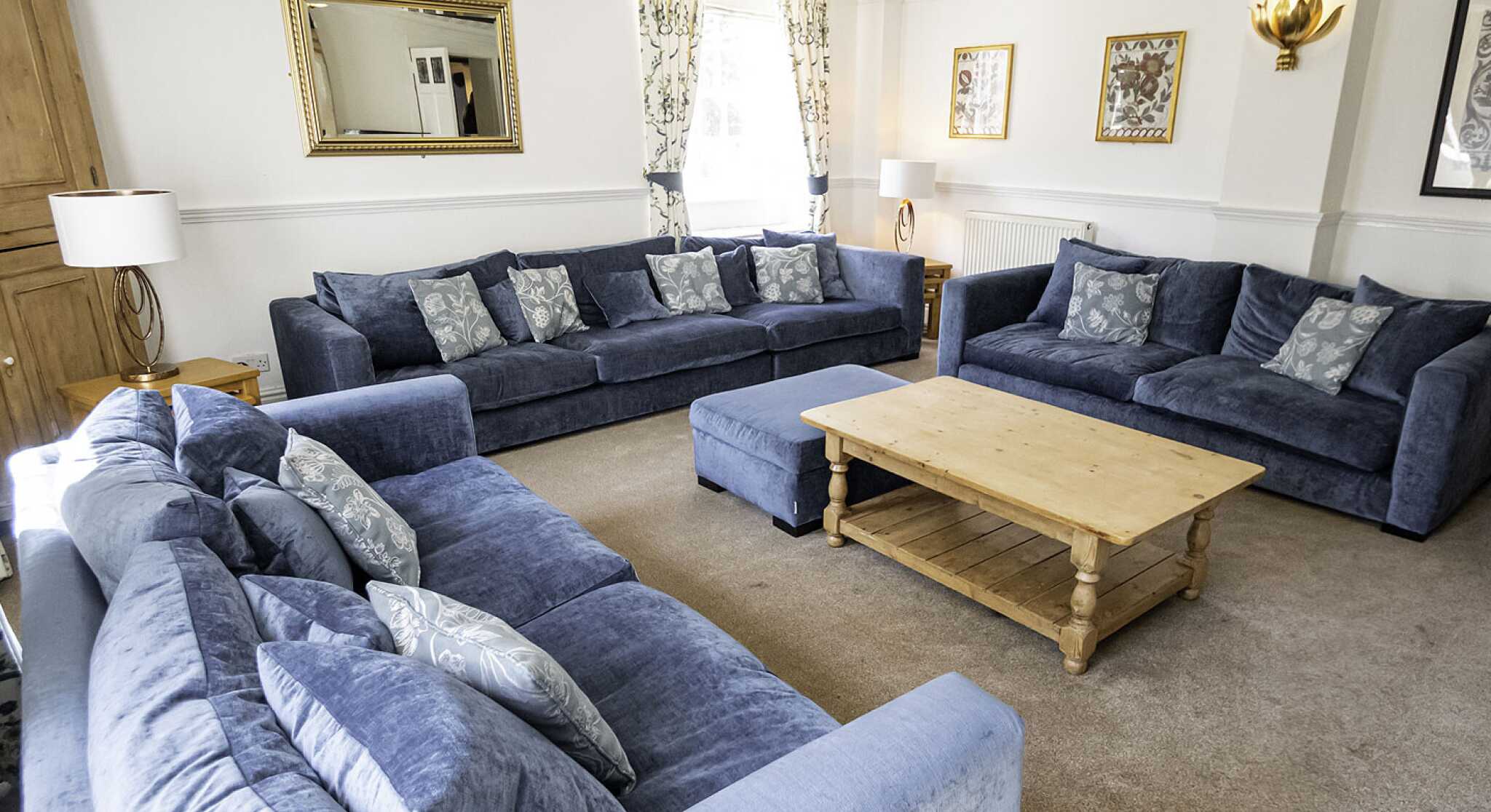 A spacious living room featuring two large blue sofas and an ottoman, all adorned with decorative cushions. There's a wooden coffee table in the center, surrounded by light-colored carpet. Wall decorations include a mirror and framed art, with natural light streaming in through large windows.