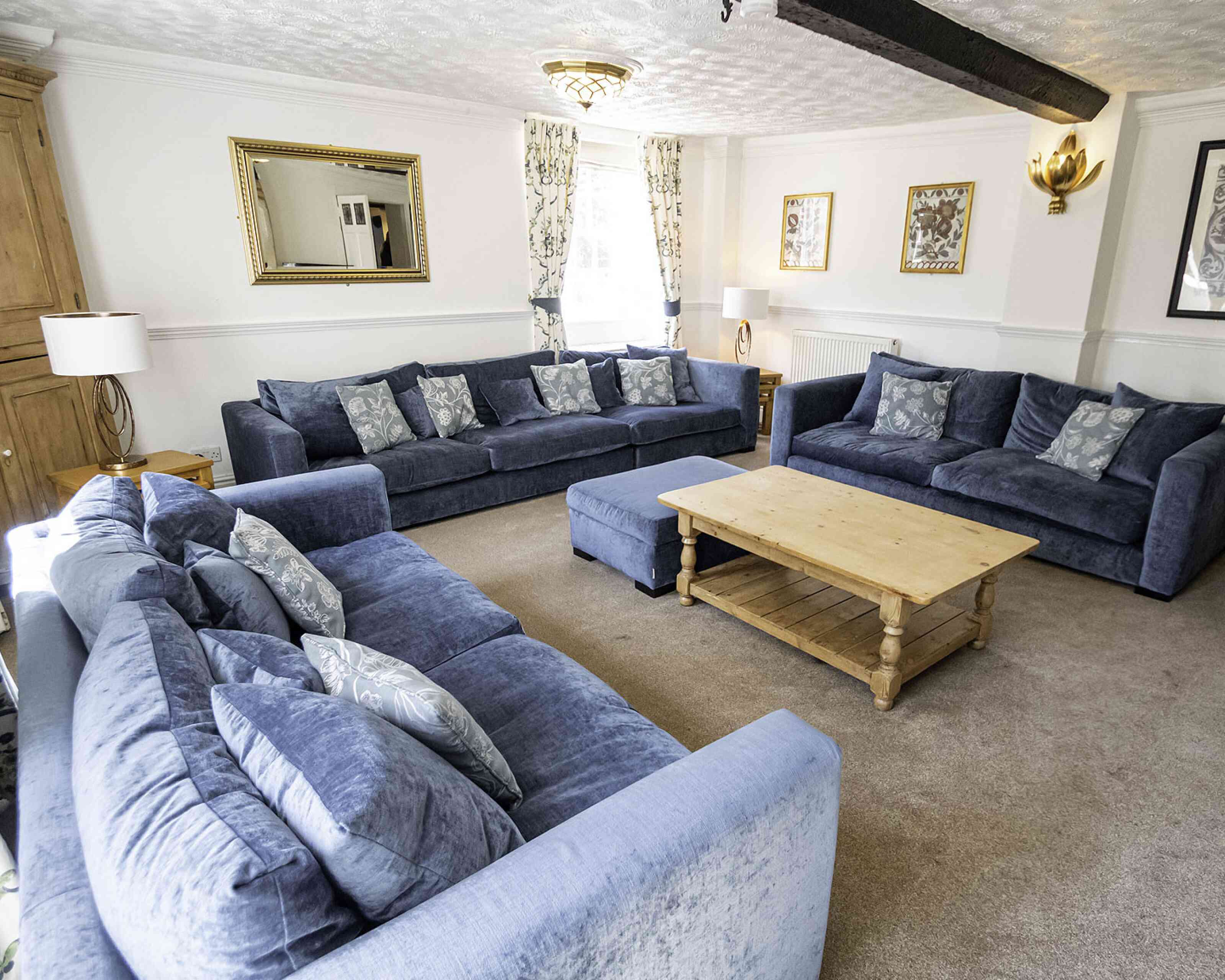 A spacious living room featuring two large blue sofas and an ottoman, all adorned with decorative cushions. There's a wooden coffee table in the center, surrounded by light-colored carpet. Wall decorations include a mirror and framed art, with natural light streaming in through large windows.
