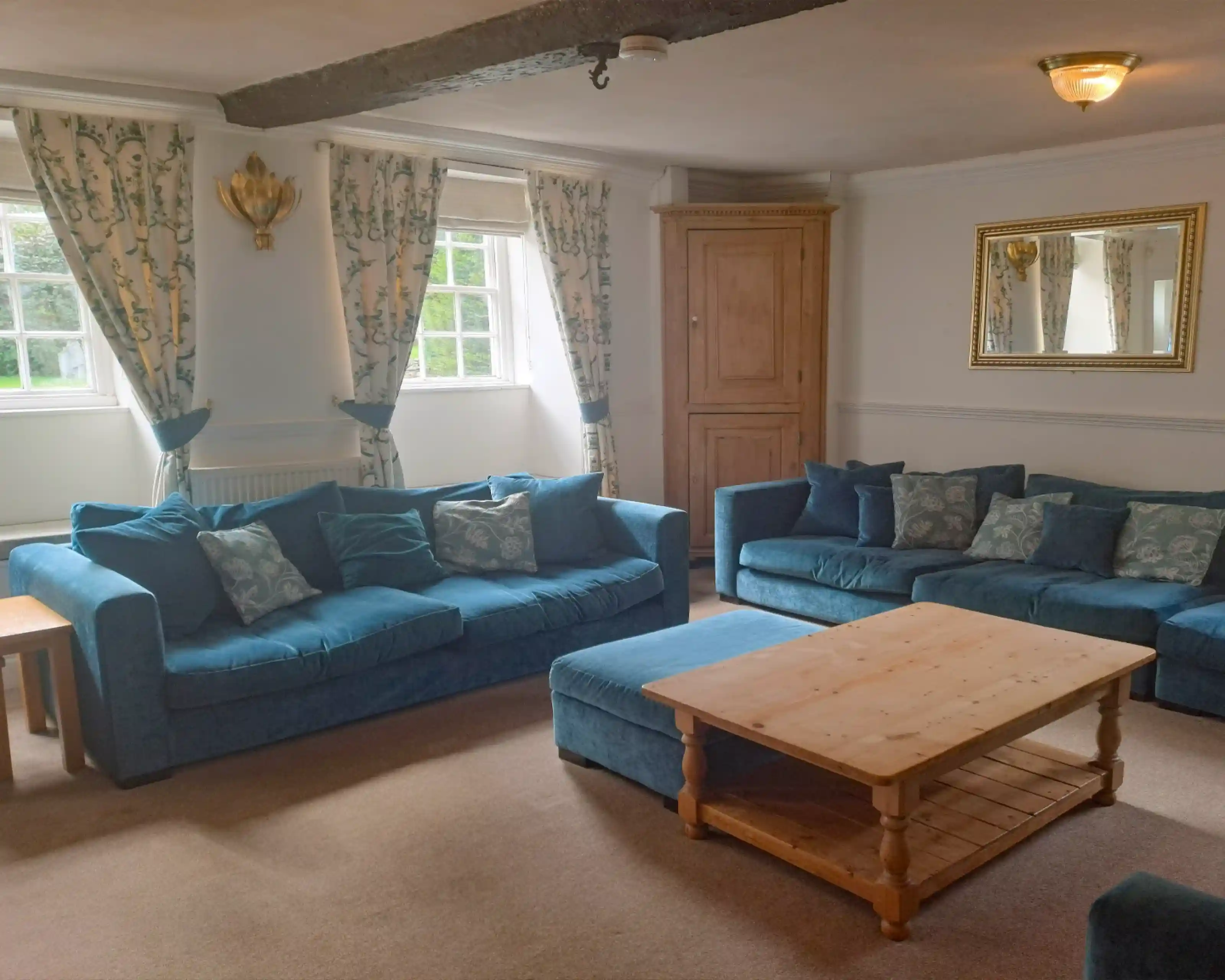 A cozy living room featuring two blue sofas with decorative cushions, a light wooden coffee table in the center, and large windows dressed with floral curtains. A mirror hangs on the wall, and there's a wooden cabinet in the corner.