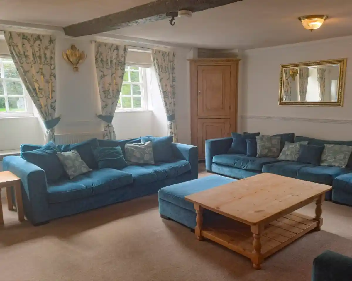 A cozy living room featuring two blue sofas with decorative cushions, a light wooden coffee table in the center, and large windows dressed with floral curtains. A mirror hangs on the wall, and there's a wooden cabinet in the corner.