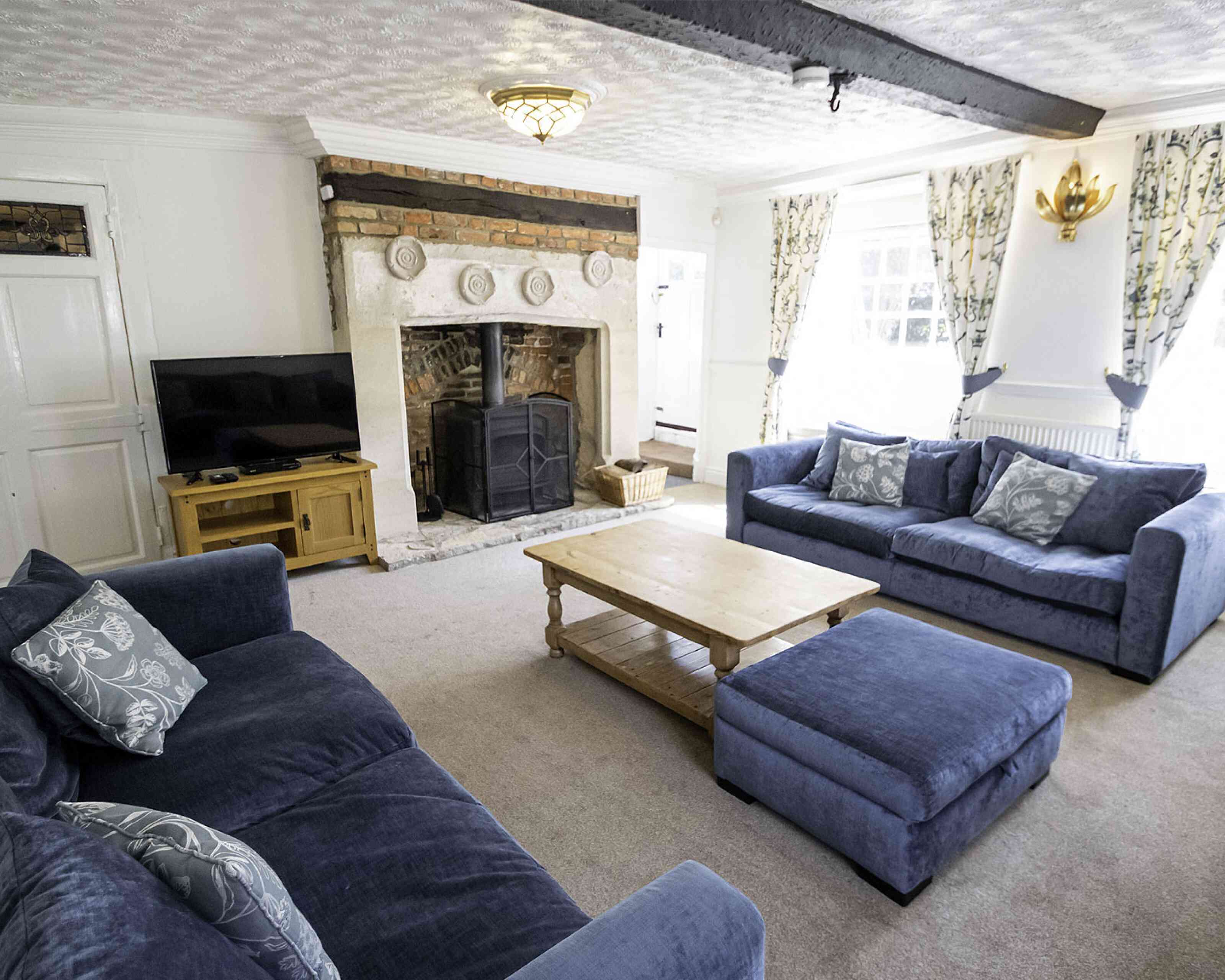 Cozy living room featuring two blue sofas and a matching ottoman. A wooden coffee table sits in the center, and a stone fireplace with a log burner is set into the wall. Natural light filters through patterned curtains.