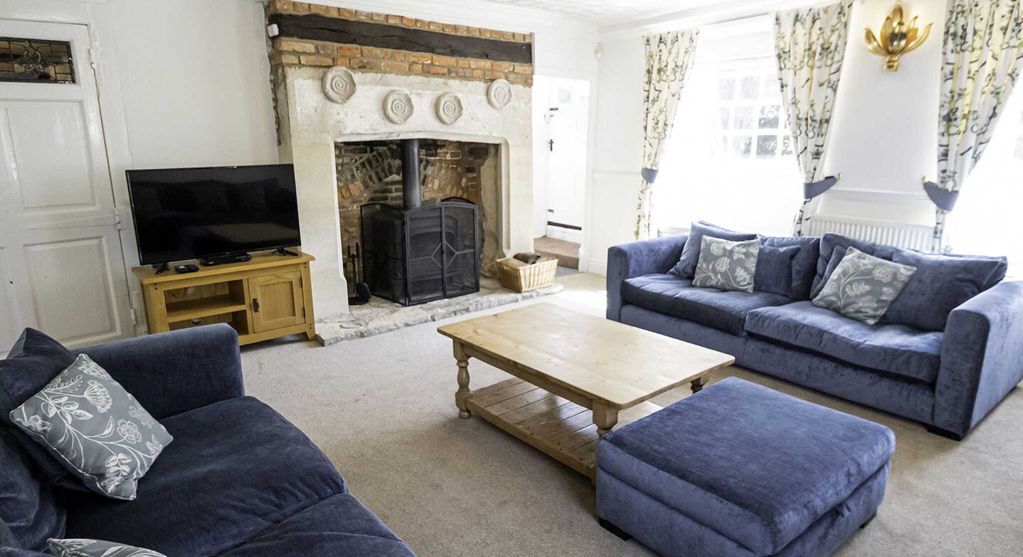 Cozy living room featuring two blue sofas and a matching ottoman. A wooden coffee table sits in the center, and a stone fireplace with a log burner is set into the wall. Natural light filters through patterned curtains.