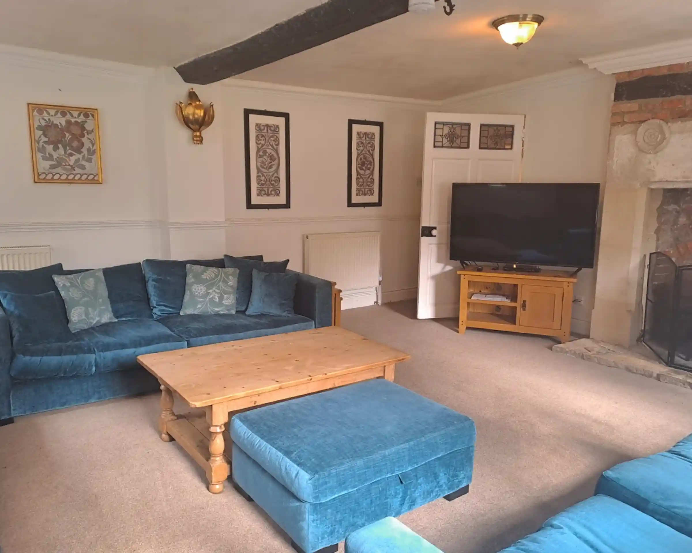A cozy living room featuring a blue plush sofa set with decorative cushions, a wooden coffee table, and a flat-screen TV on a wooden stand. The walls are adorned with framed artwork, and a fireplace is visible in the corner.