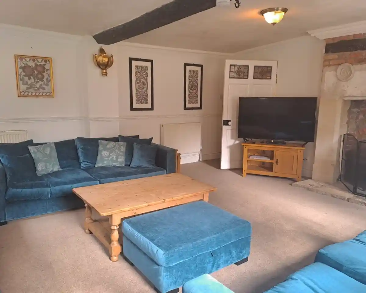 A cozy living room featuring a blue plush sofa set with decorative cushions, a wooden coffee table, and a flat-screen TV on a wooden stand. The walls are adorned with framed artwork, and a fireplace is visible in the corner.