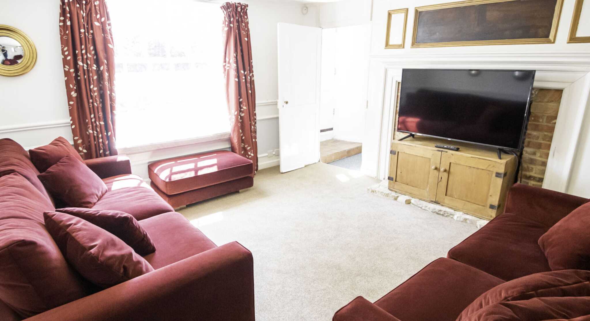 A cozy living room featuring two large red sofas, a flat-screen TV mounted above a wooden entertainment unit, and bright natural light streaming through large windows. Decorative curtains frame the windows, and there is a warm, inviting atmosphere.