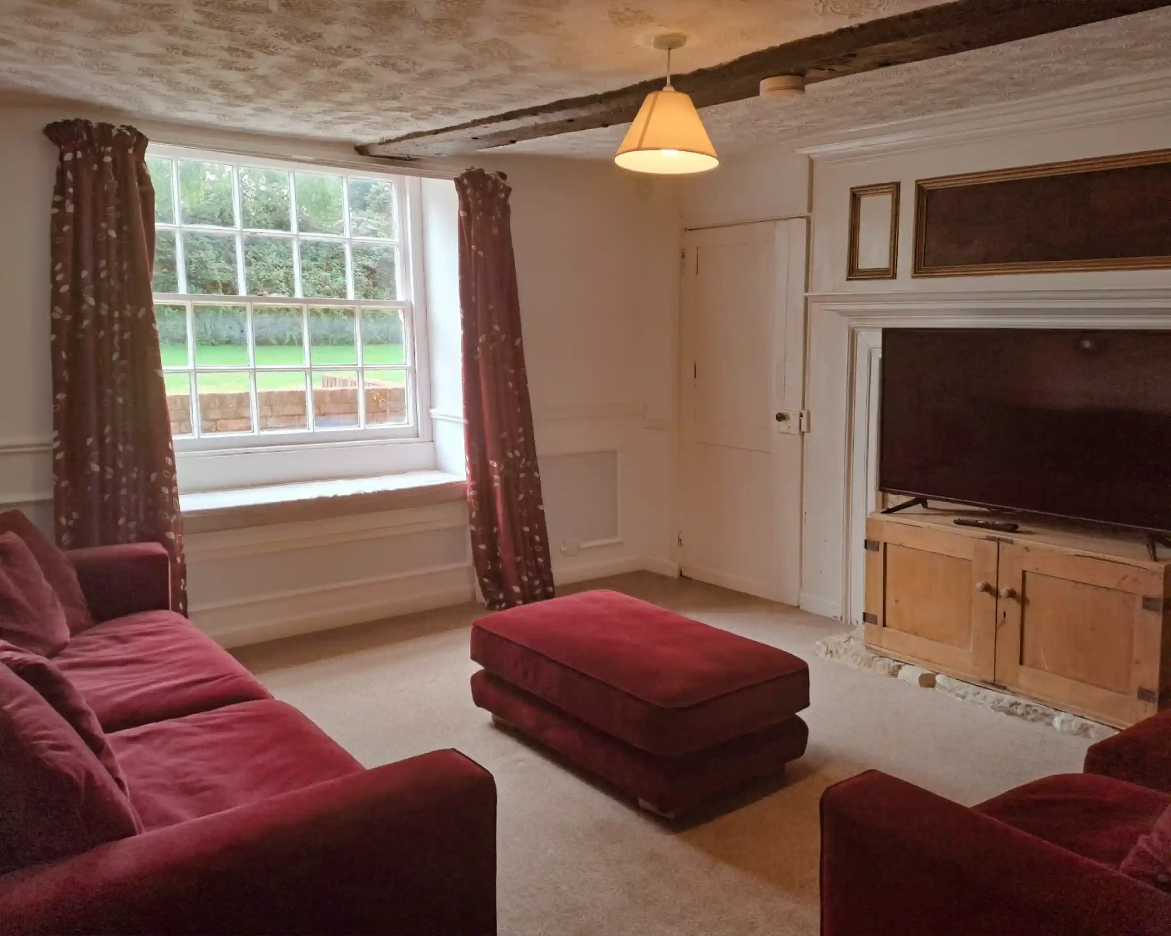 A cozy living room features two deep red sofas and a matching ottoman. A large window allows natural light to illuminate the space, framed by patterned curtains. A wooden cabinet with a television sits against the wall, complementing the bright, airy atmosphere.