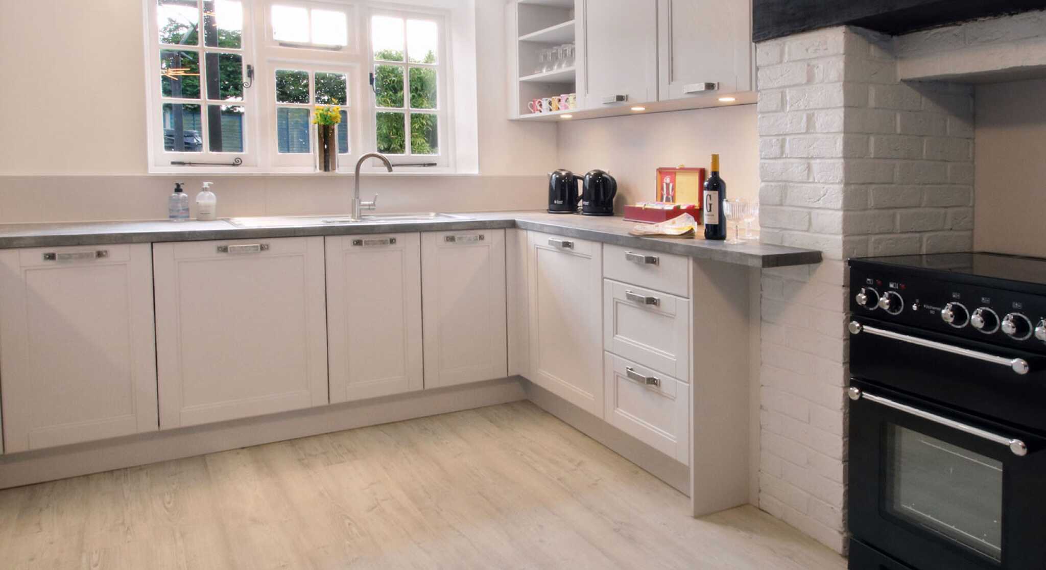 A modern, well-lit kitchen showcasing light-colored cabinetry, a stainless steel sink with a faucet, and a black stove beside a white brick oven. The space features minimalistic decor with a few utensils on display.