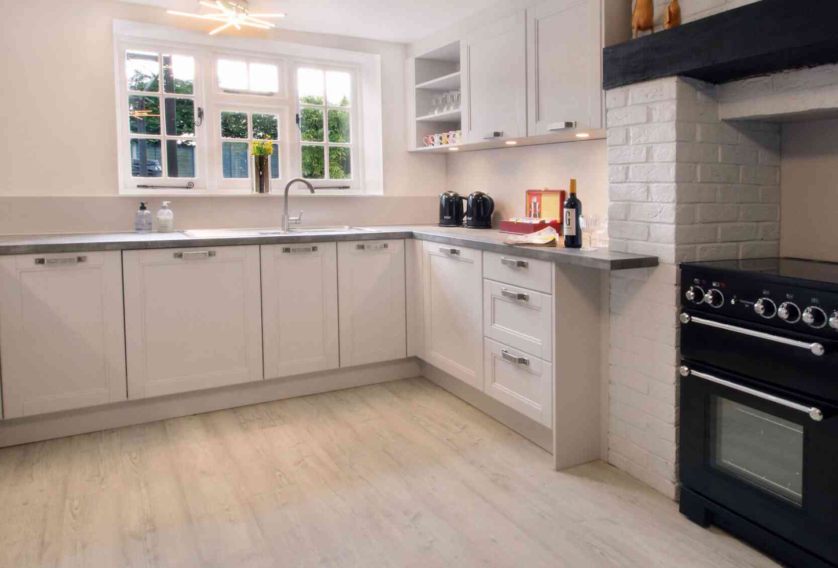 A modern, well-lit kitchen showcasing light-colored cabinetry, a stainless steel sink with a faucet, and a black stove beside a white brick oven. The space features minimalistic decor with a few utensils on display.
