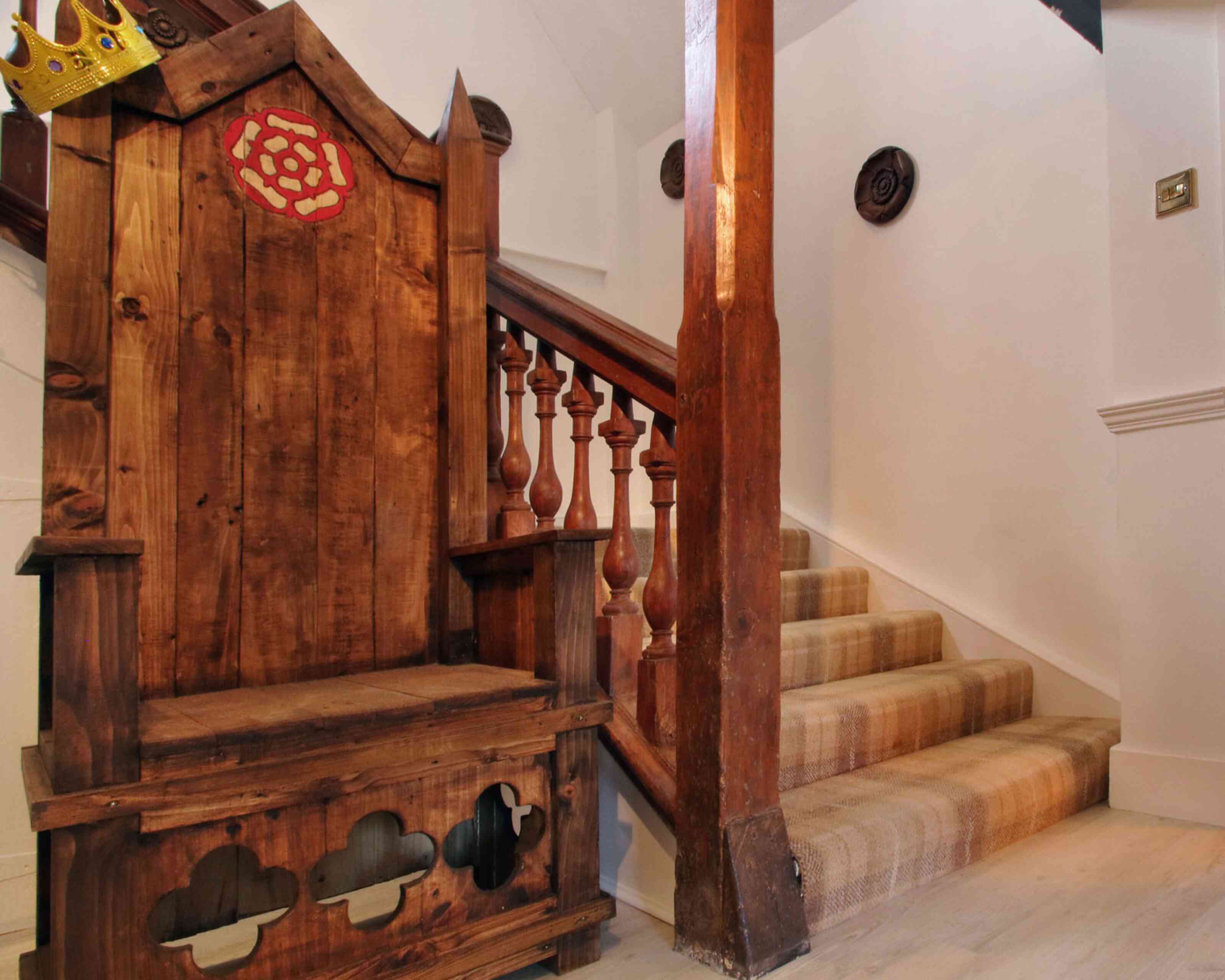 A wooden throne with a red emblem sits next to a staircase with wooden balusters. The staircase is carpeted and leads to an upper level, with white walls and decorative features.