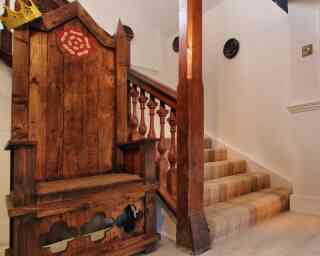A wooden throne with a red emblem sits next to a staircase with wooden balusters. The staircase is carpeted and leads to an upper level, with white walls and decorative features.