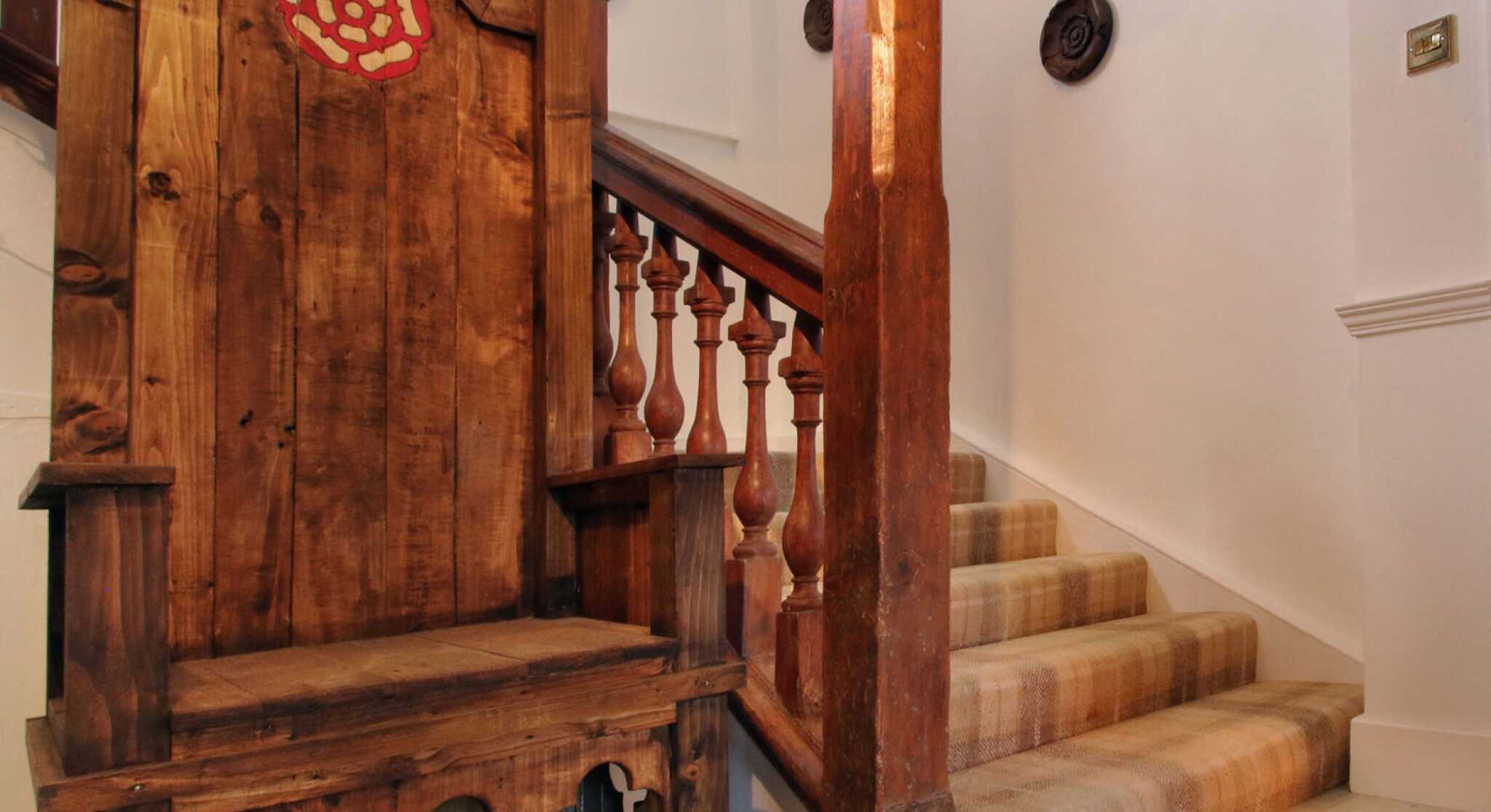 A wooden throne with a red emblem sits next to a staircase with wooden balusters. The staircase is carpeted and leads to an upper level, with white walls and decorative features.