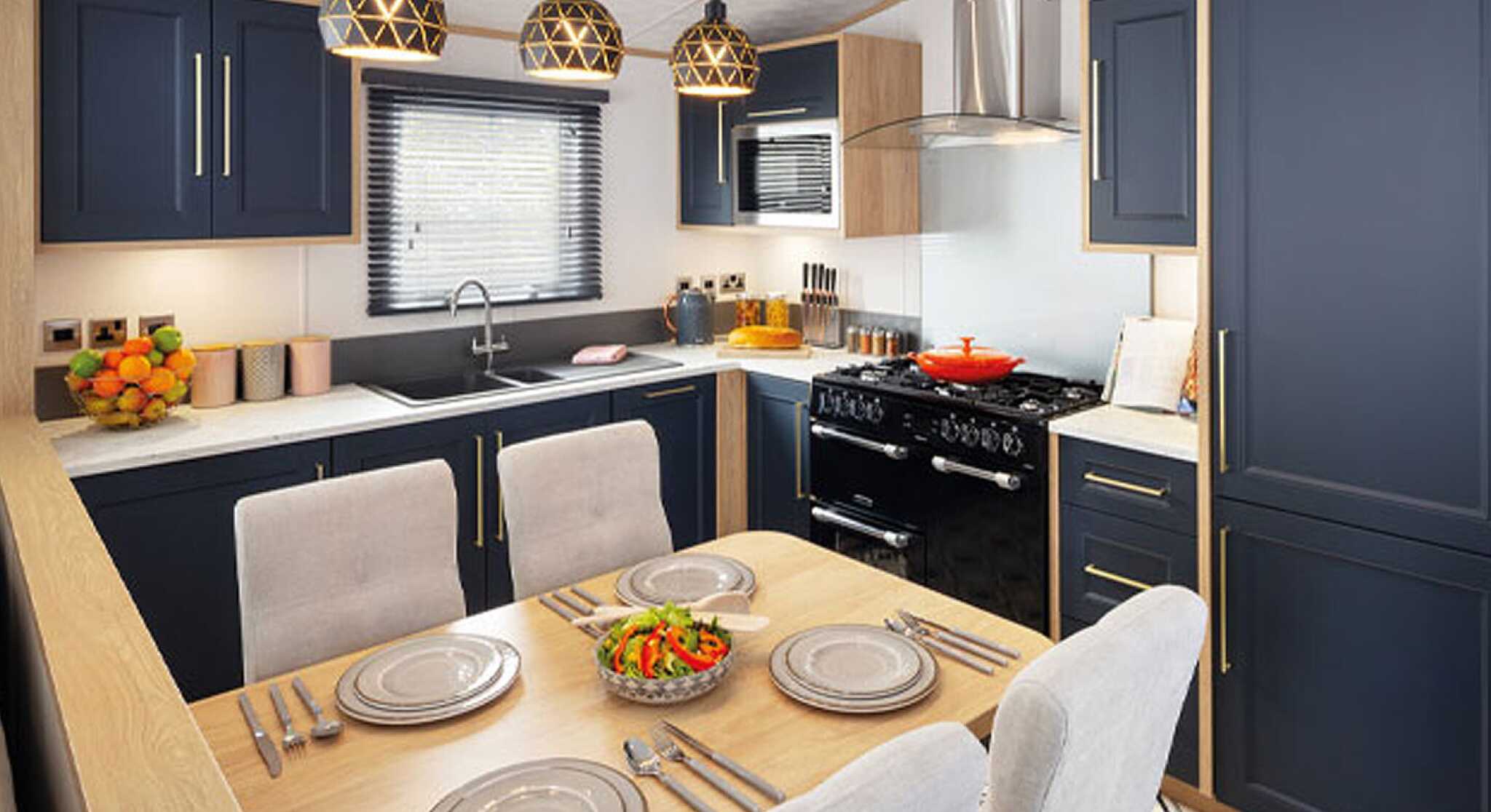 A modern kitchen featuring navy blue cabinetry, a wooden countertop, and a dining area with a table set for four. Pendant lights hang above the table, and a black stove is visible in the background. Fresh salad is placed in the center of the table against a patterned floor.