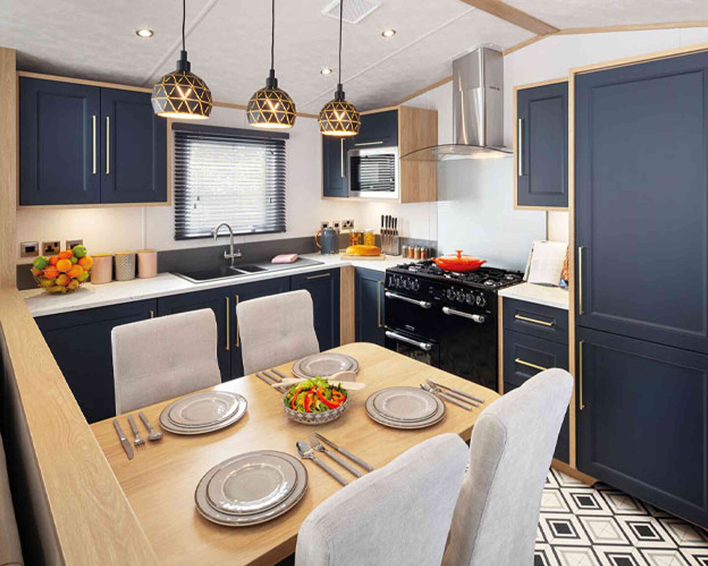 A modern kitchen featuring navy blue cabinetry, a wooden countertop, and a dining area with a table set for four. Pendant lights hang above the table, and a black stove is visible in the background. Fresh salad is placed in the center of the table against a patterned floor.