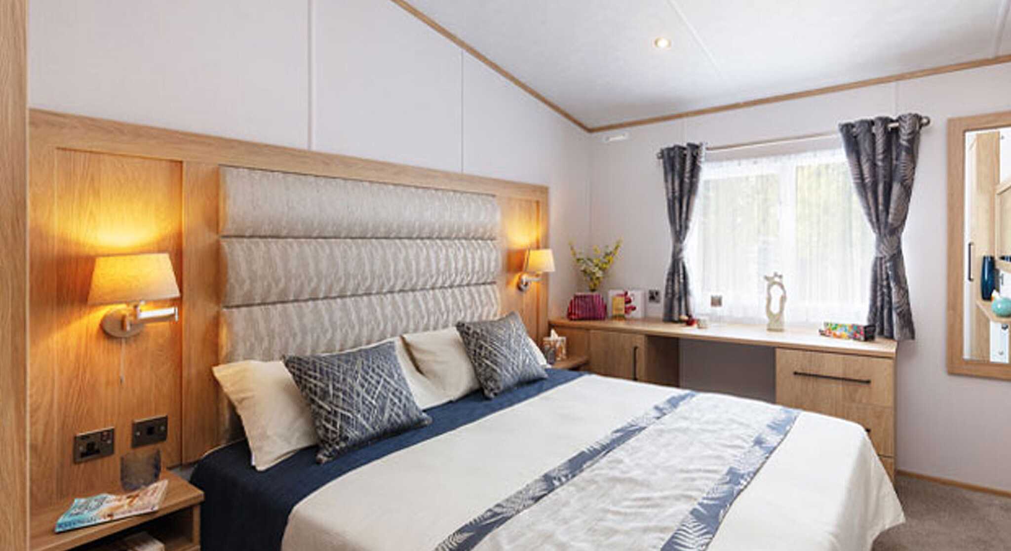 A cozy bedroom with a large bed featuring a padded headboard, decorative pillows, and a soft bedspread. Wood accents surround the room, including bedside tables with lamps and a dresser. Natural light filters through a window with sheer curtains.