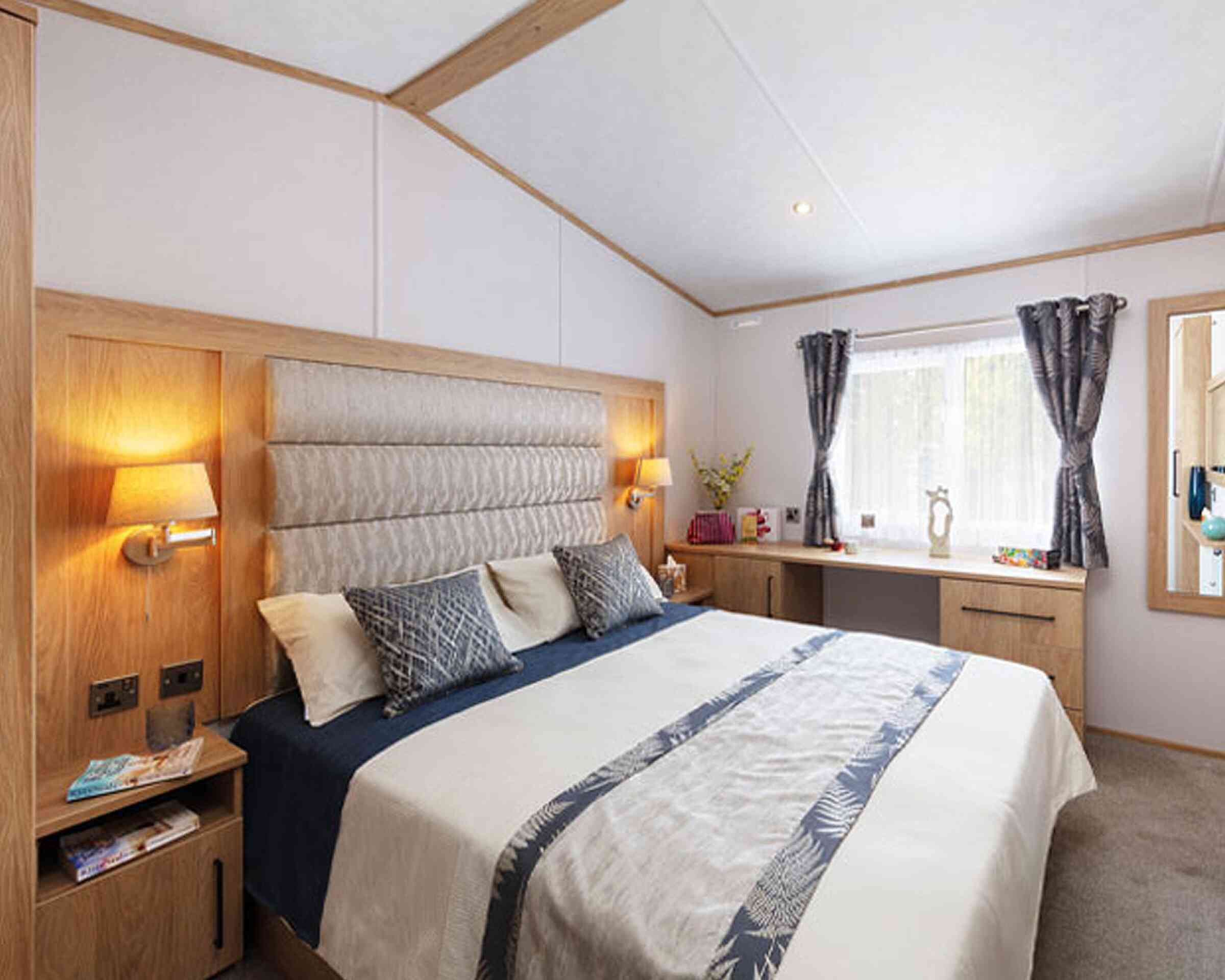A cozy bedroom with a large bed featuring a padded headboard, decorative pillows, and a soft bedspread. Wood accents surround the room, including bedside tables with lamps and a dresser. Natural light filters through a window with sheer curtains.