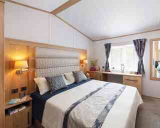 A cozy bedroom with a large bed featuring a padded headboard, decorative pillows, and a soft bedspread. Wood accents surround the room, including bedside tables with lamps and a dresser. Natural light filters through a window with sheer curtains.