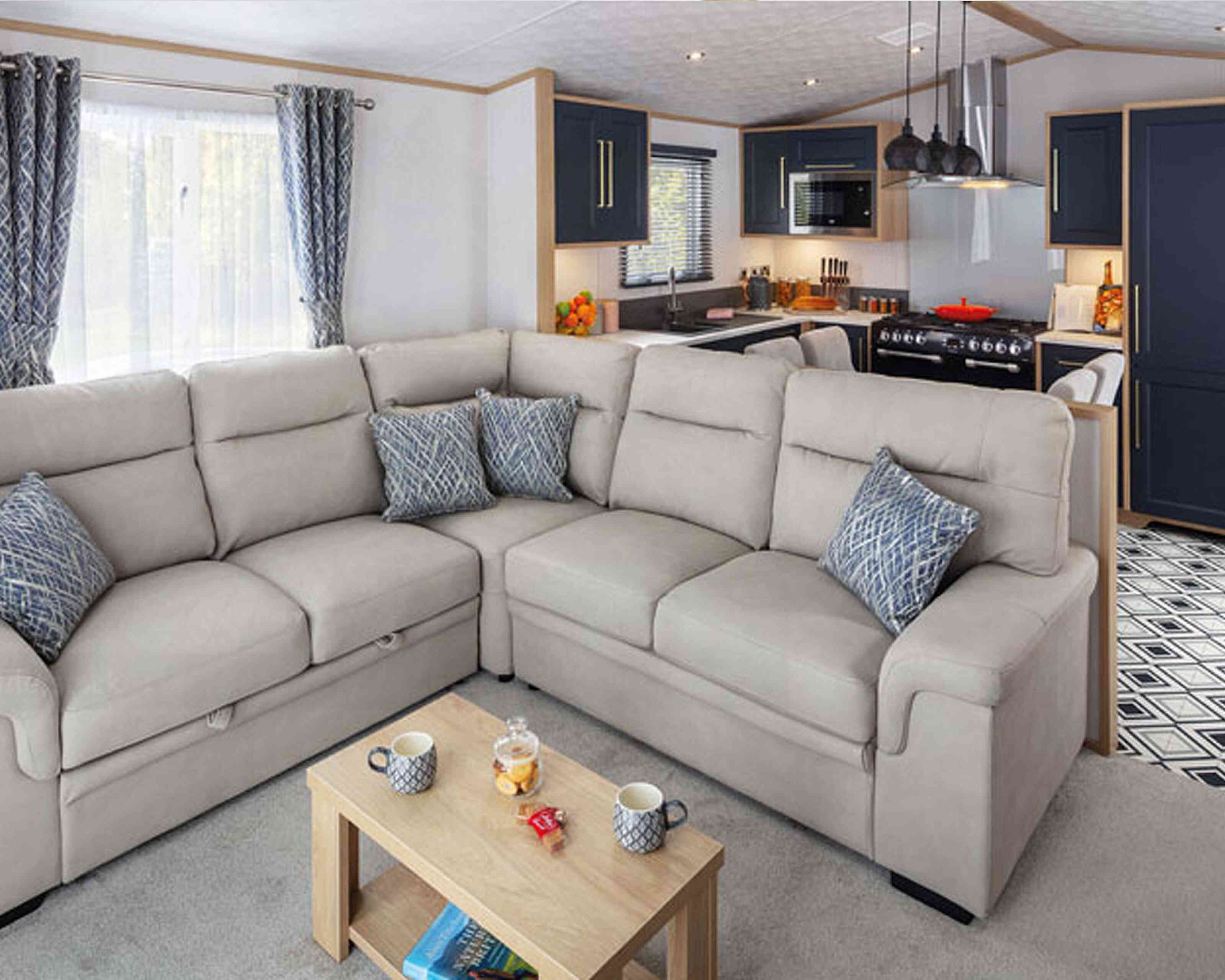 Cozy living area featuring a light-colored sectional sofa with decorative pillows, a wooden coffee table with snacks, and a modern kitchen in the background with dark cabinets and appliances.