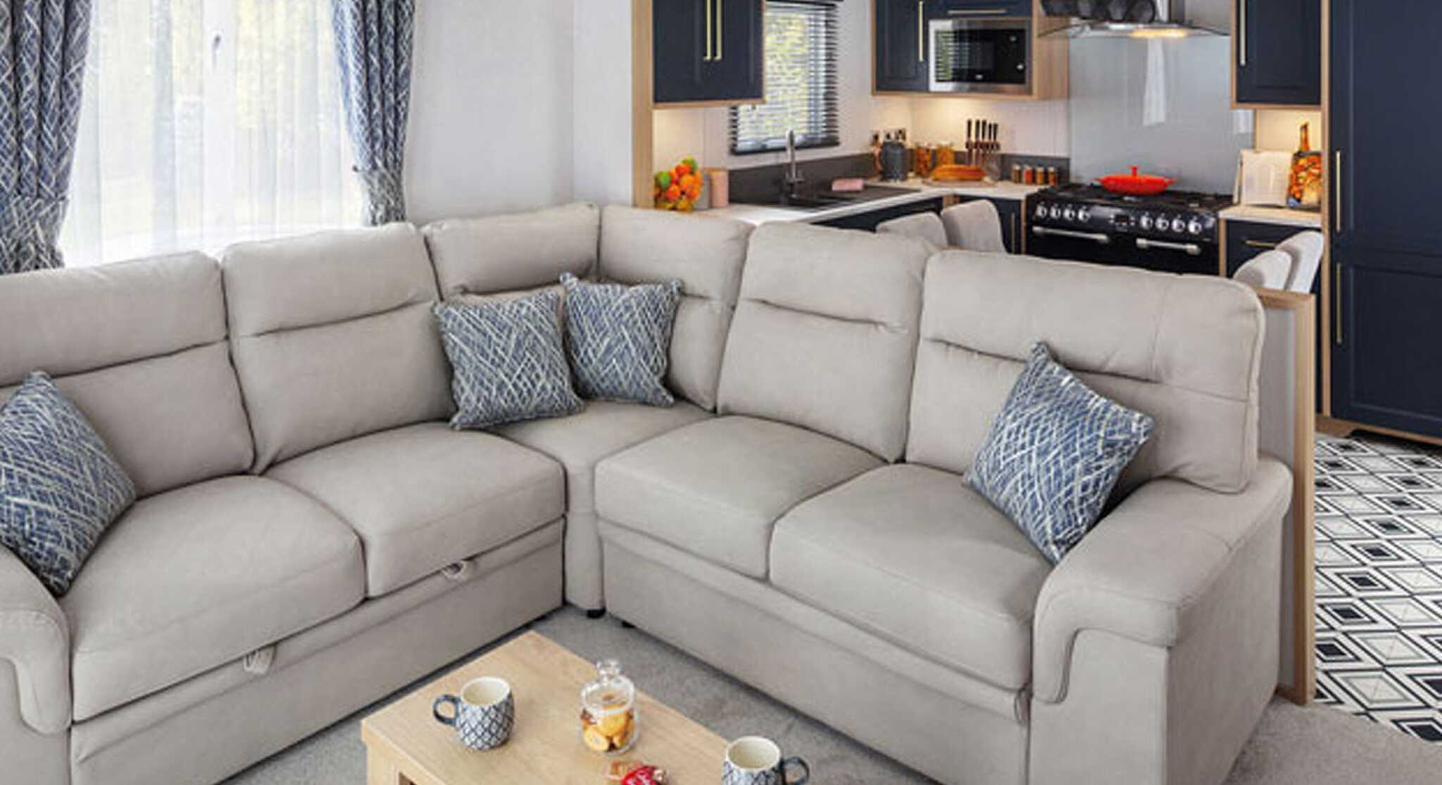 Cozy living area featuring a light-colored sectional sofa with decorative pillows, a wooden coffee table with snacks, and a modern kitchen in the background with dark cabinets and appliances.