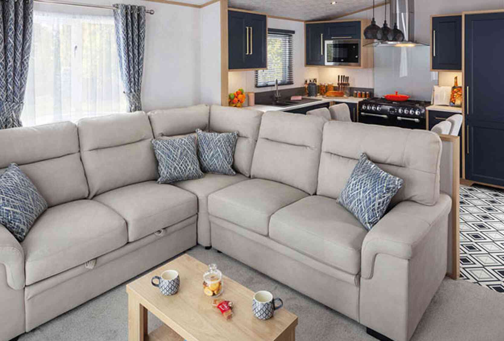 Cozy living area featuring a light-colored sectional sofa with decorative pillows, a wooden coffee table with snacks, and a modern kitchen in the background with dark cabinets and appliances.