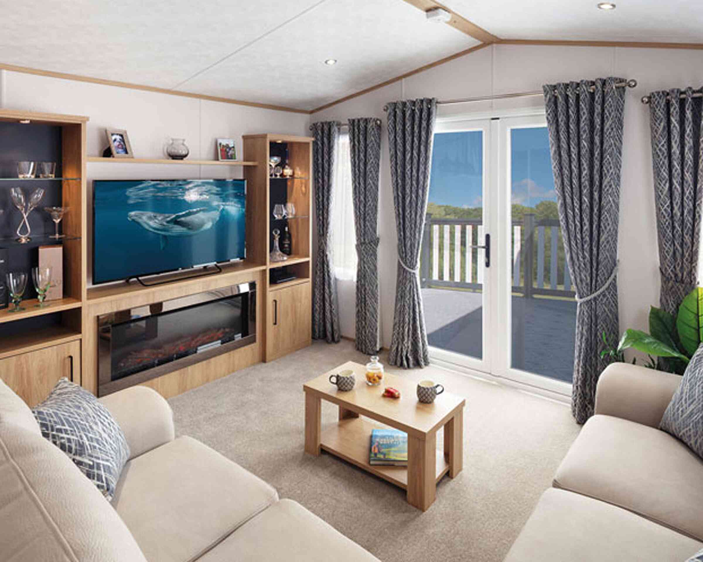 A cozy living room featuring a light-colored sofa, a modern entertainment unit with a television displaying a marine scene, and a coffee table with snacks. Large curtains frame the window that leads to a balcony, with a view of greenery outside. Soft lighting adds warmth to the space.
