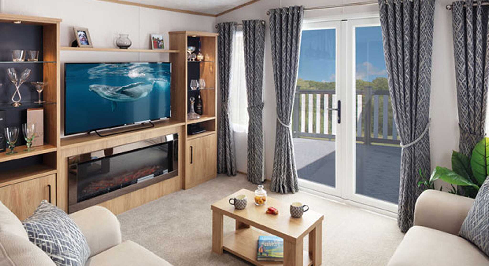 A cozy living room featuring a light-colored sofa, a modern entertainment unit with a television displaying a marine scene, and a coffee table with snacks. Large curtains frame the window that leads to a balcony, with a view of greenery outside. Soft lighting adds warmth to the space.