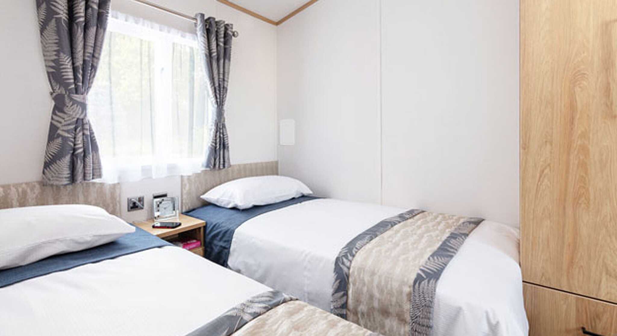 A cozy bedroom with two single beds dressed in white linens and blue accents. The room features light wood paneling, a window with sheer curtains, and decorative fern-patterned curtains. A bedside table holds a small lamp and clock.