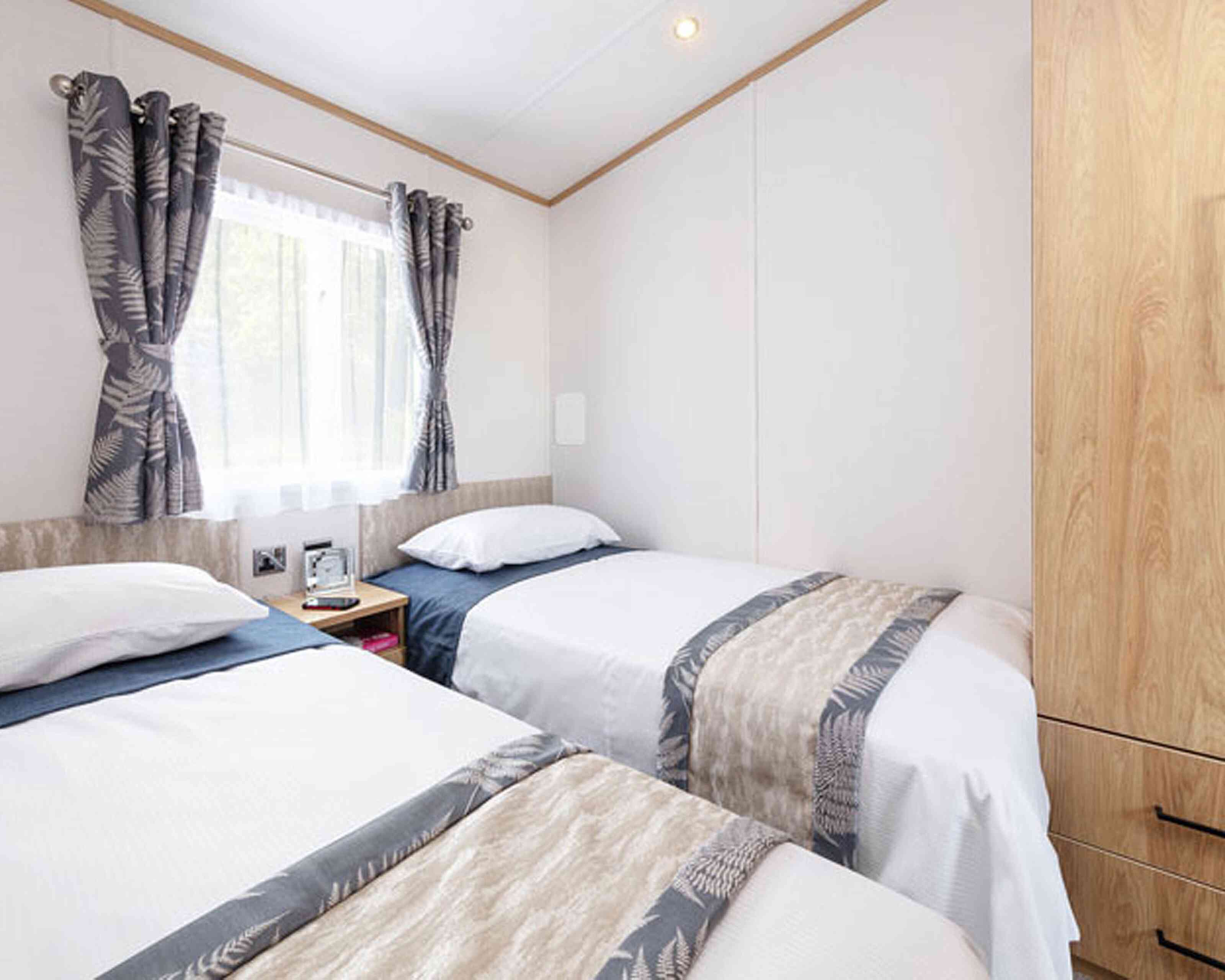 A cozy bedroom with two single beds dressed in white linens and blue accents. The room features light wood paneling, a window with sheer curtains, and decorative fern-patterned curtains. A bedside table holds a small lamp and clock.
