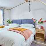 A cozy bedroom featuring a double bed with light bedding, flanked by a wooden nightstand. A warm orange throw and a yellow folded blanket are draped over the bed. There are fresh flowers in a vase and a small cup on the nightstand, along with a decorative basket nearby. The room has soft natural light coming through a window with checkered curtains.