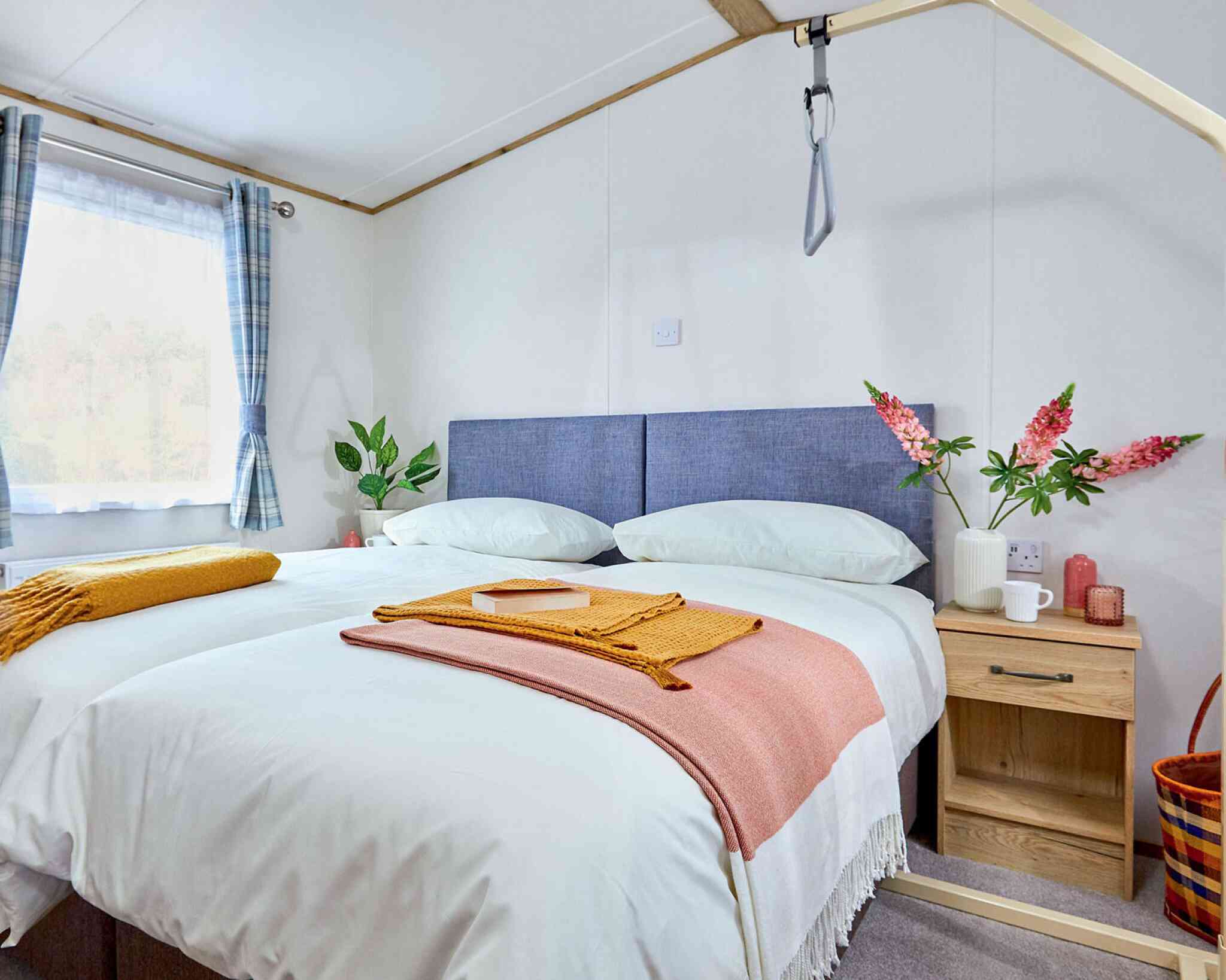 A cozy bedroom featuring a double bed with light bedding, flanked by a wooden nightstand. A warm orange throw and a yellow folded blanket are draped over the bed. There are fresh flowers in a vase and a small cup on the nightstand, along with a decorative basket nearby. The room has soft natural light coming through a window with checkered curtains.