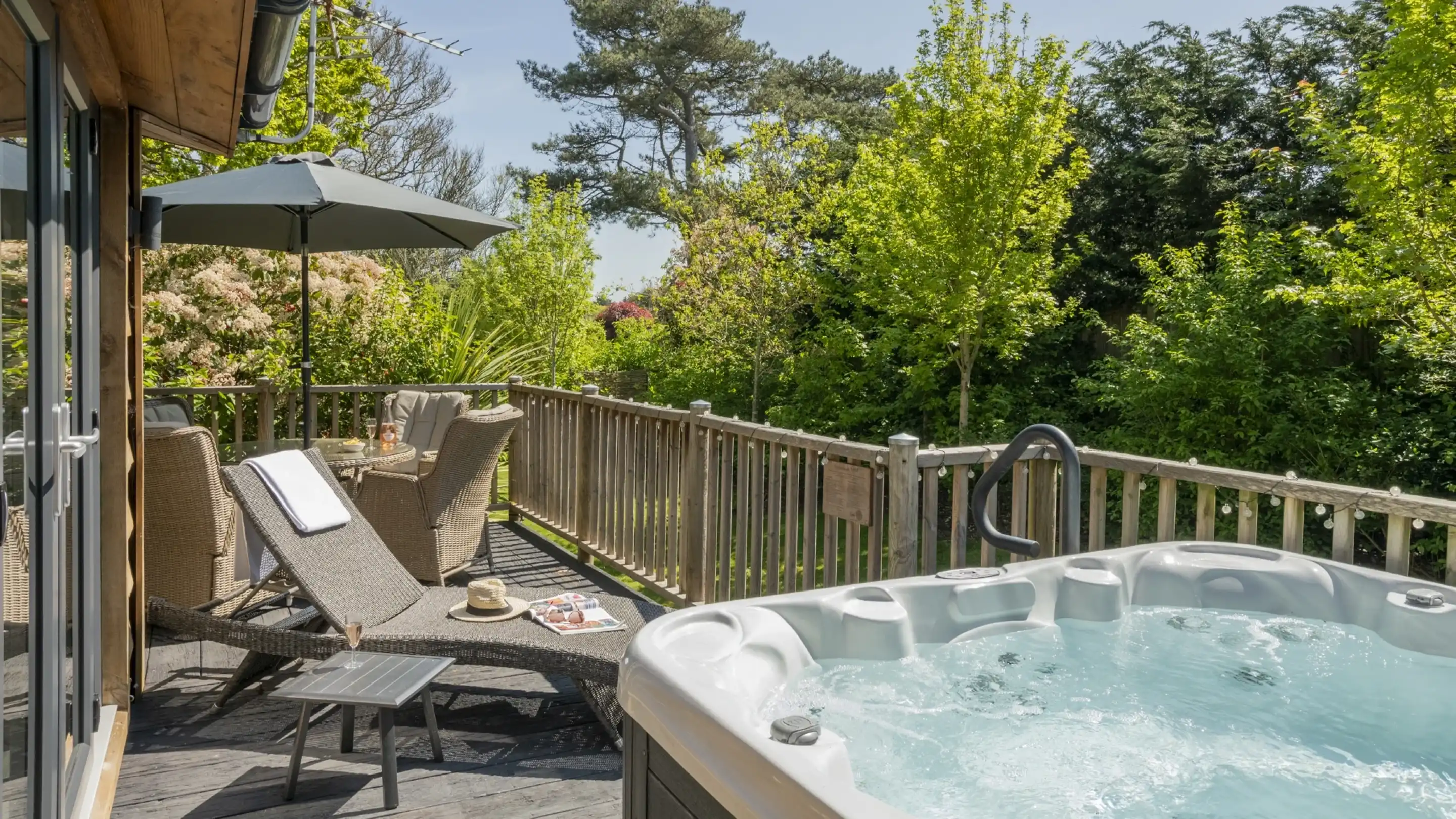 A tranquil outdoor deck features a hot tub, a shaded seating area with comfortable chairs, and a lounge chair. Lush greenery surrounds the space, brightened by sunlight filtering through the trees.